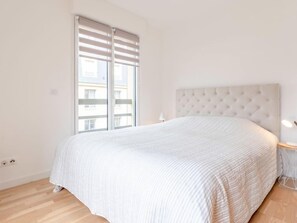A bedroom with a large bed, a window with blinds, a tufted headboard, and a bedside table with a lamp.
