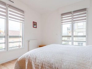 Een slaapkamer met een groot bed, twee ramen met luxaflex, een radiator en een klein houten nachtkastje.