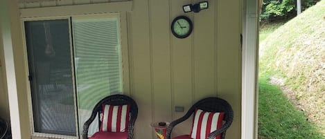 Un porche cubierto con dos sillas de mimbre con cojines rojos, un reloj y un jarrón decorativo.