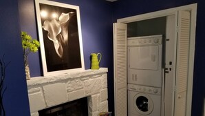 A room with a white brick fireplace, a framed abstract painting, a green pitcher, and a stacked washer and dryer.
