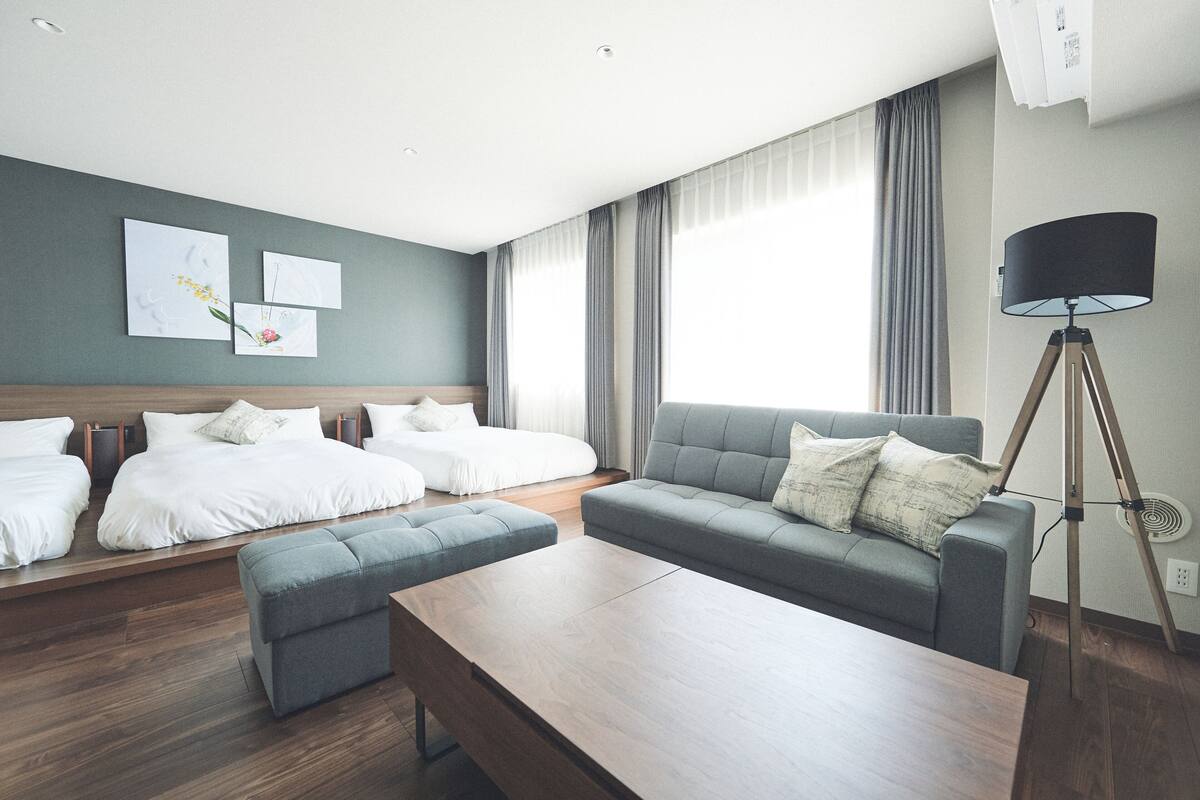 Une chambre d’hôtel moderne avec deux lits, un canapé gris, une table basse en bois et un lampadaire.