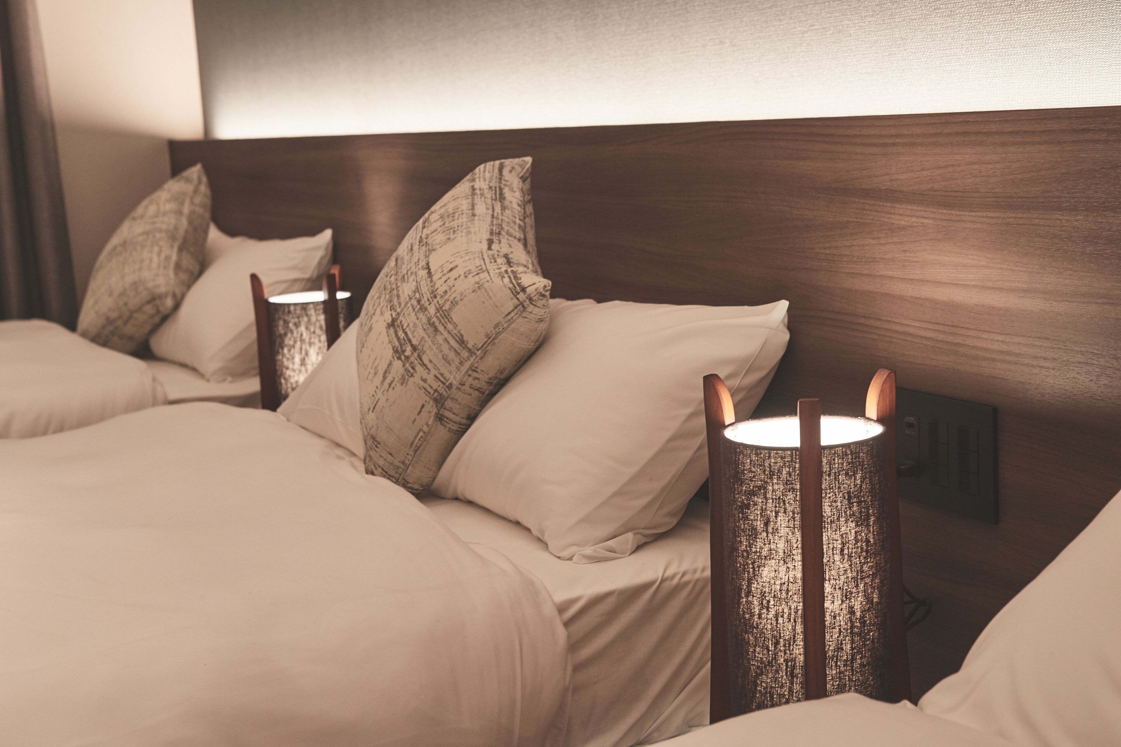 A hotel room with a wooden headboard, two bedside tables with lamps, and a bed with beige bedding and patterned pillows.