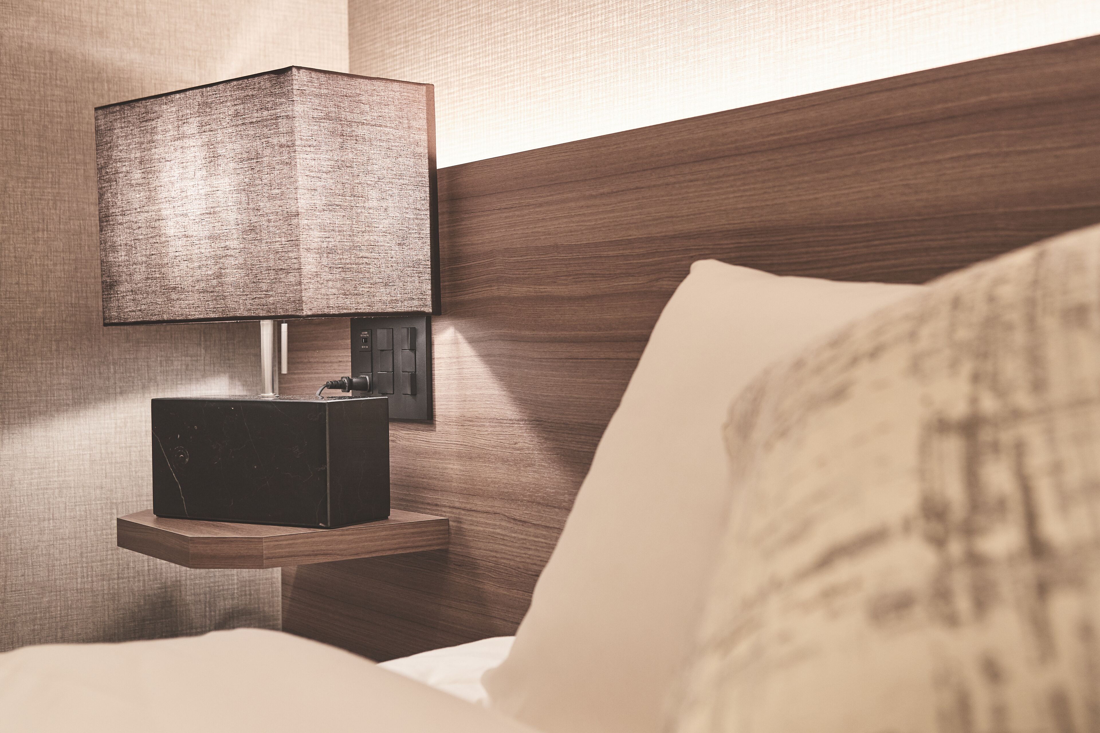 A neatly made bed with a wooden headboard, a bedside table with a lamp, and a power outlet.