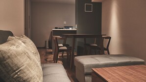 A modern living room with a sofa, ottoman, and wooden coffee table. A dining area with chairs and a table is visible in the background. - ReLA Higashimatsudo (Matsudo)