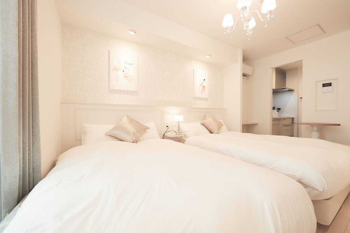 Une chambre d’hôtel avec deux lits, une table de chevet avec une lampe, un climatiseur fixé au mur et un petit coin cuisine.