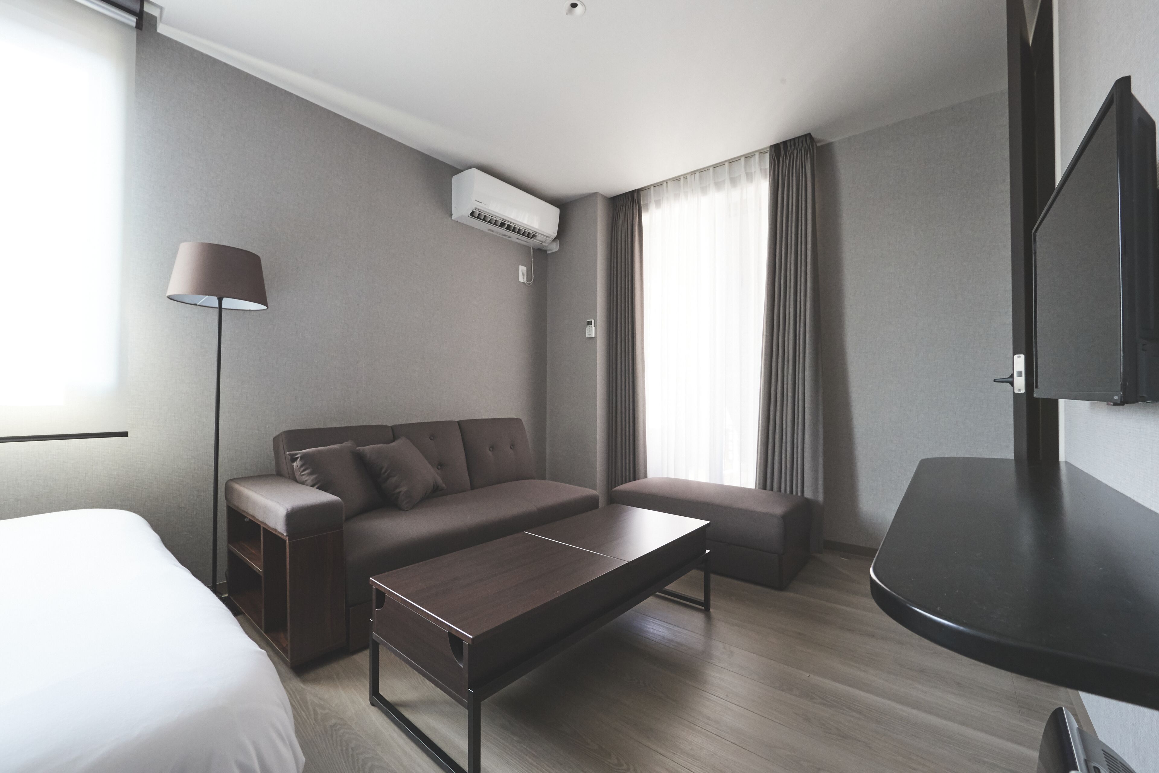A modern hotel room with a bed, a sofa, a small table, a lamp, an air conditioning unit, and a TV.