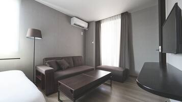 A modern hotel room with a bed, a sofa, a small table, a lamp, an air conditioning unit, and a TV.
