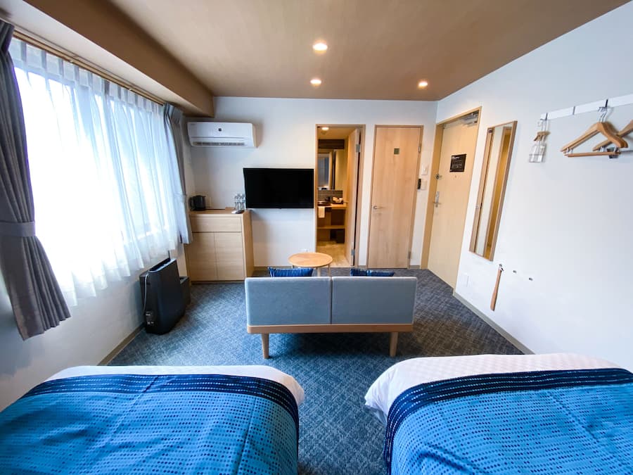 A hotel room with two beds, a desk, a TV, and a bathroom visible through an open door.