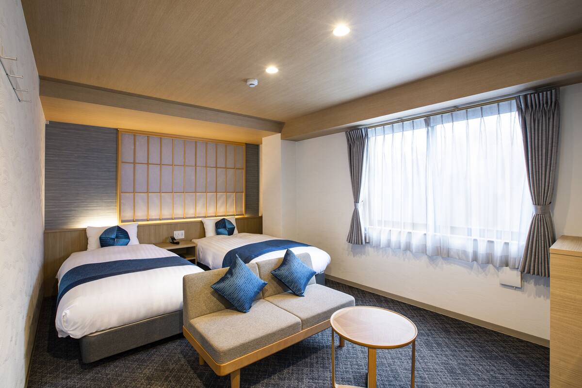 A hotel room with two beds, a sofa, a small table, and a large window with curtains.