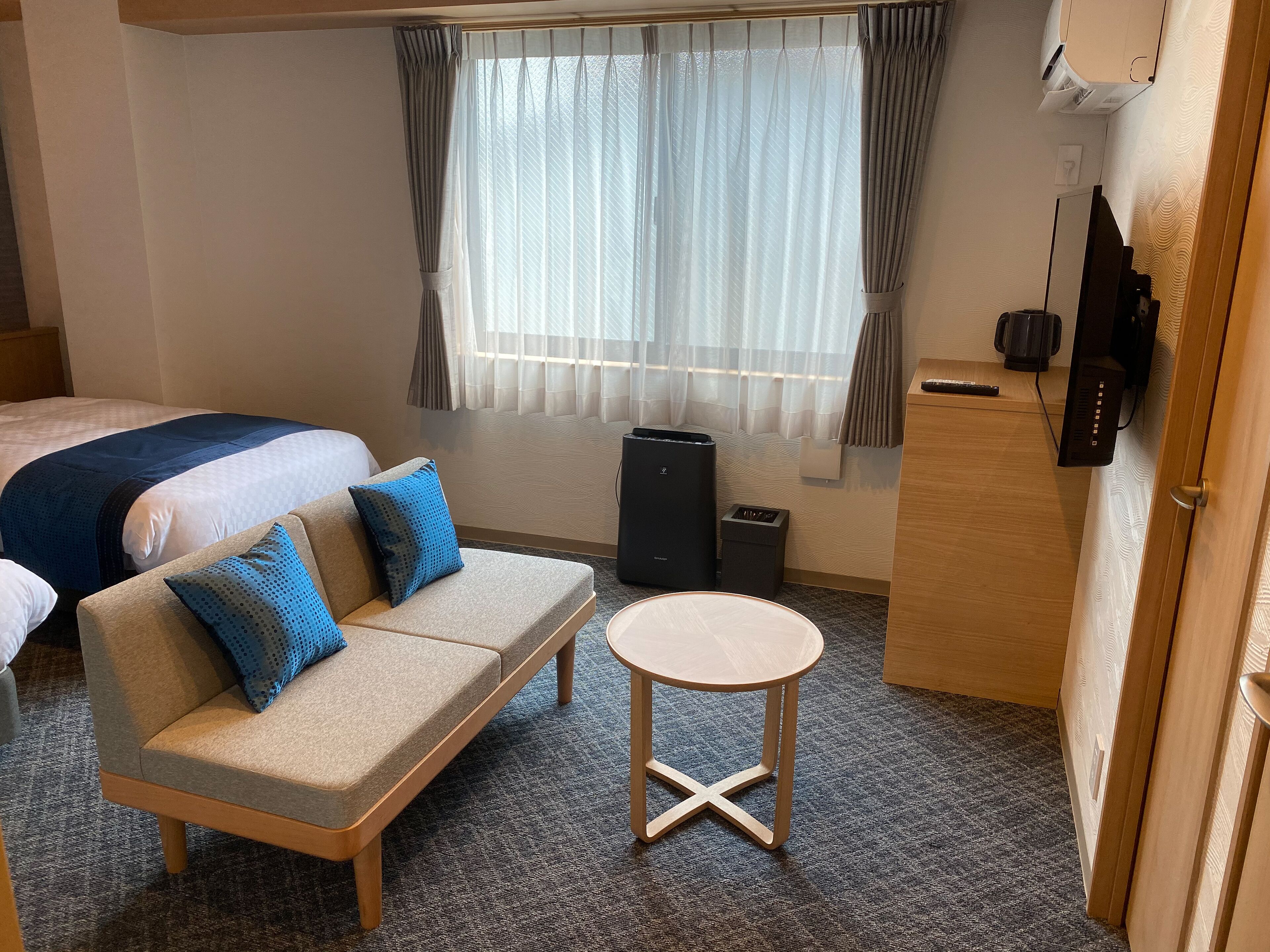 A hotel room with a bed, a sofa with blue pillows, a small round table, and a TV on a wooden cabinet.