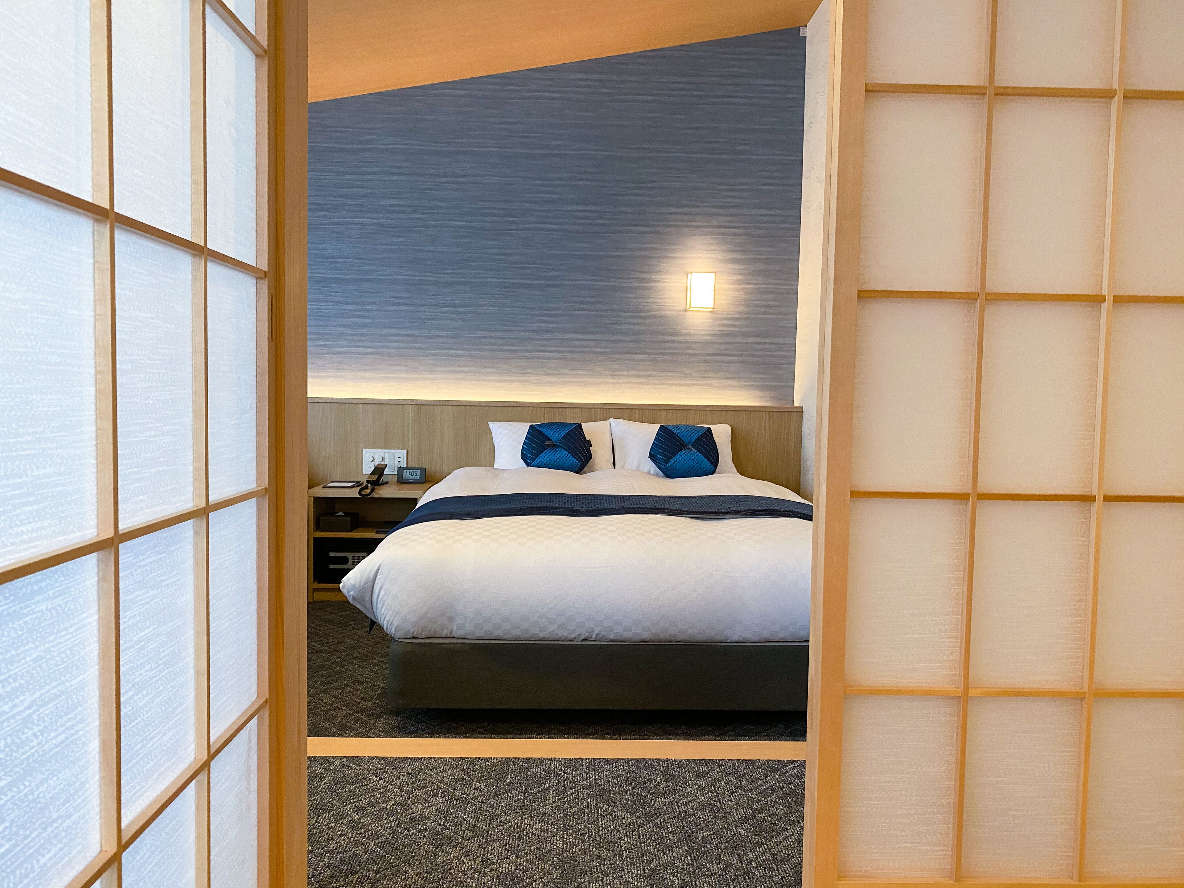 A hotel room with a bed, a nightstand, and a wall-mounted light.