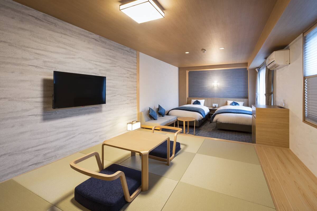 A modern hotel room with a flat-screen TV, a sofa, a bed with blue bedding, a small table, and tatami mats on the floor.