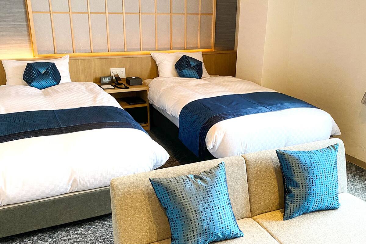 A hotel room with two beds, a sofa, a nightstand with a lamp, and a window with blinds.