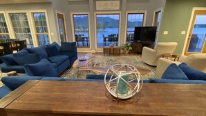 A spacious living room with a large wooden dining table, blue sofas, and a view of the outdoors through large windows. - The Fun Spot 3 - Serene Home with Beautiful Lake & Mtn Views to Relax and Enjoy! (Huddleston)