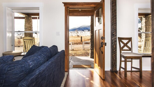 A room with a wooden floor, a blue sofa, a wooden chair, and a door leading to an outdoor area with a view of mountains.
