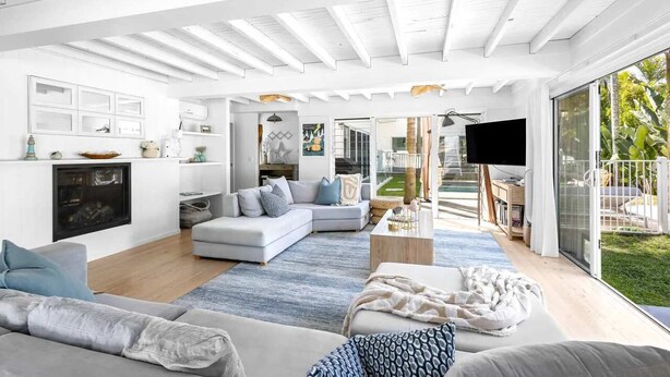 A spacious living area with a large sofa, a coffee table, and a TV. There is a fireplace, a kitchen area, and a view of the outdoors. - Barra Luxe Beach House 4BR Yaroomba (Point Arkwright)