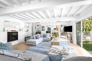 A spacious living area with a large sofa, a coffee table, and a TV. There is a fireplace, a kitchen area, and a view of the outdoors.