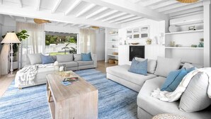 A spacious living room with a large sofa, a coffee table, and a TV. There is a bookshelf with decorative items and a view of the outdoors. - Barra Luxe Beach House 4BR Yaroomba (Point Arkwright)