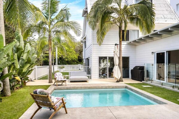 A backyard with a swimming pool, palm trees, a white house, and a covered patio area.