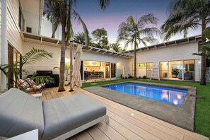 A modern outdoor pool area with a lounge chair, palm trees, and a covered patio.