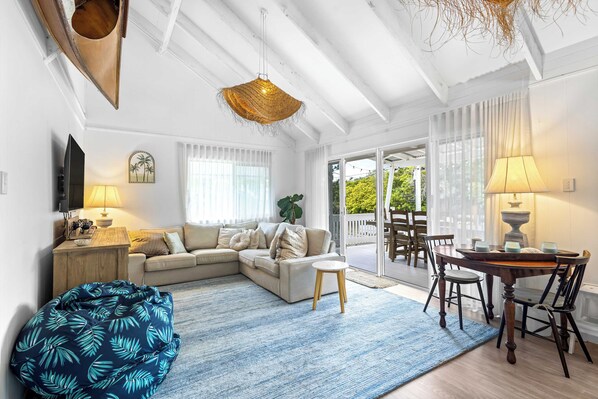 A modern living room with a sectional sofa, a coffee table, a blue bean bag, a dining table with chairs, and a large window with curtains.