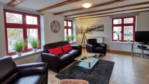 Una sala de estar con sofá, sillones, una mesa de centro y televisor. Se ven ventanas con marcos rojos y plantas en los alféizares.