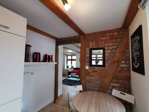 A compact kitchen with white cabinets, a wooden ceiling, and a brick wall. There's a small dining area with a round table and a chair.