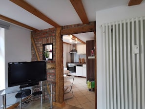 A living room with a TV, a glass table, a brick wall, and a kitchen in the background.