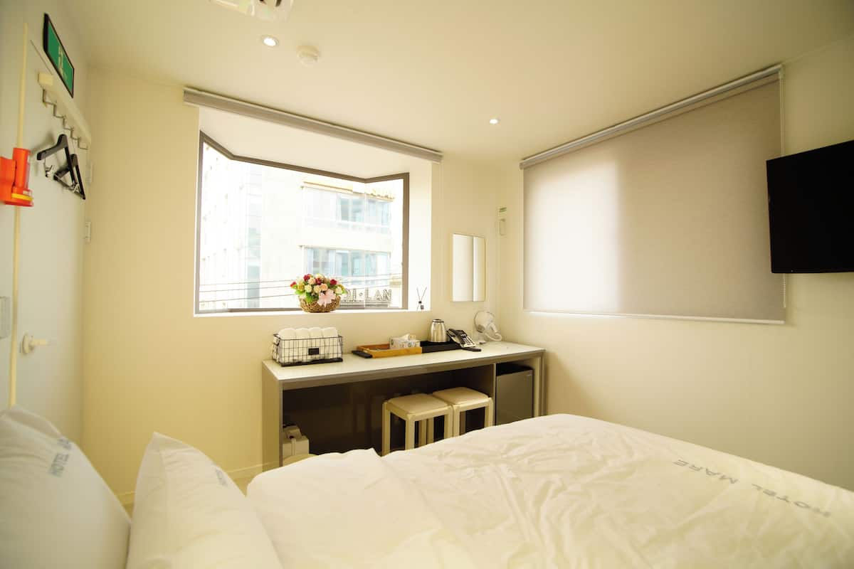 A hotel room with a bed, a desk with a telephone, a mirror, and a window with a view of buildings.