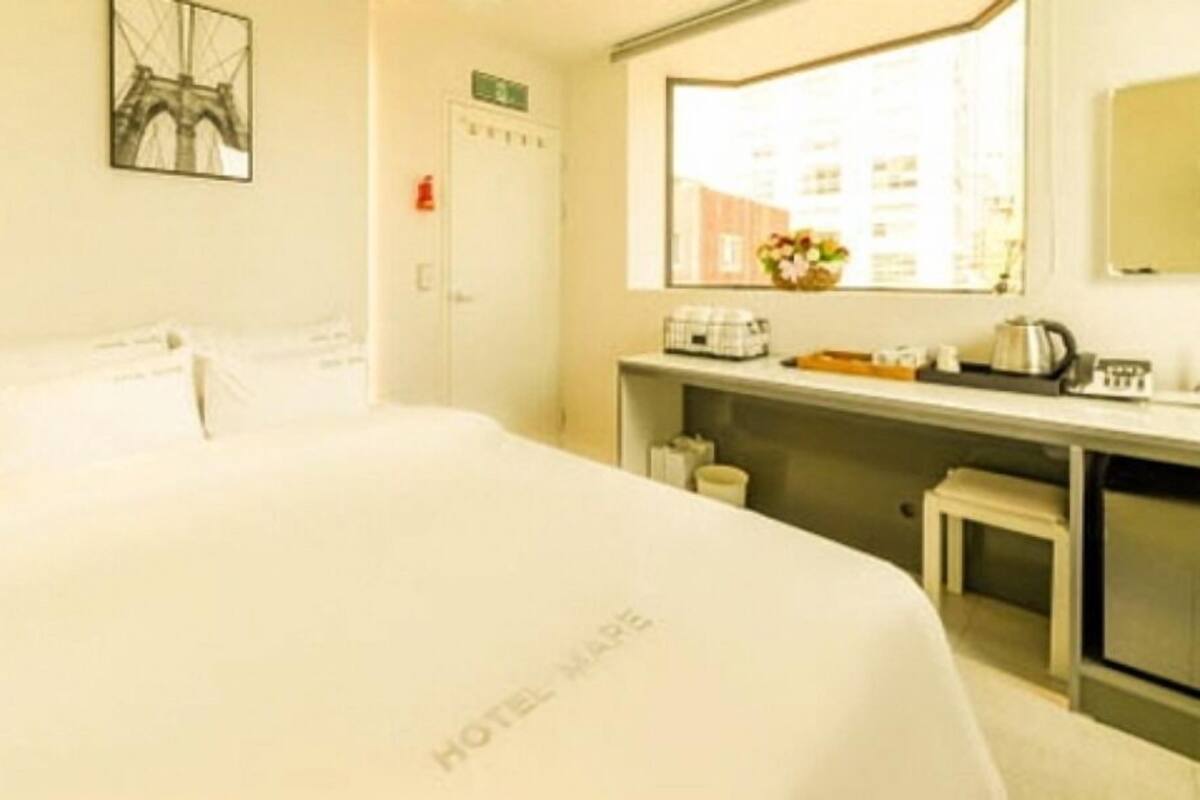 A hotel room with a bed, a desk with a kettle, a small stool, and a window with a view of buildings.