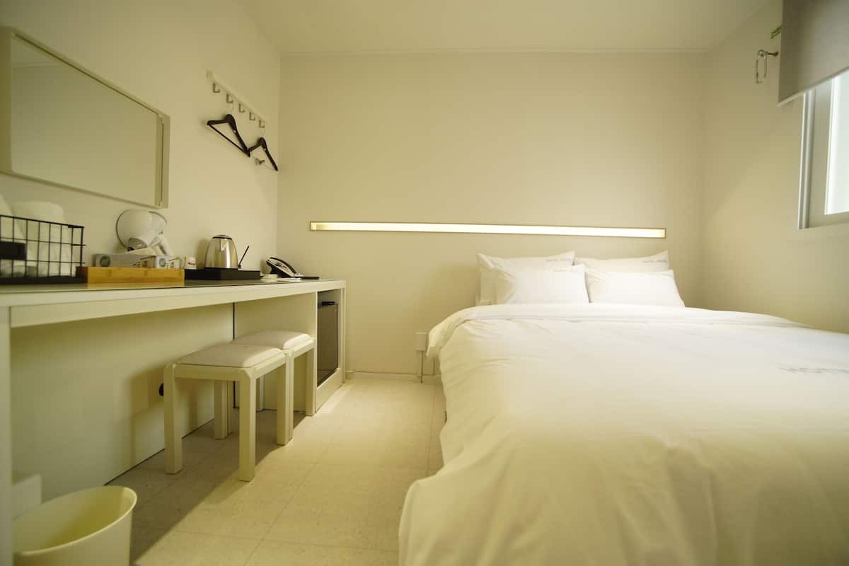A hotel room with a bed, a desk with a chair, a small stool, a trash bin, and a mirror.