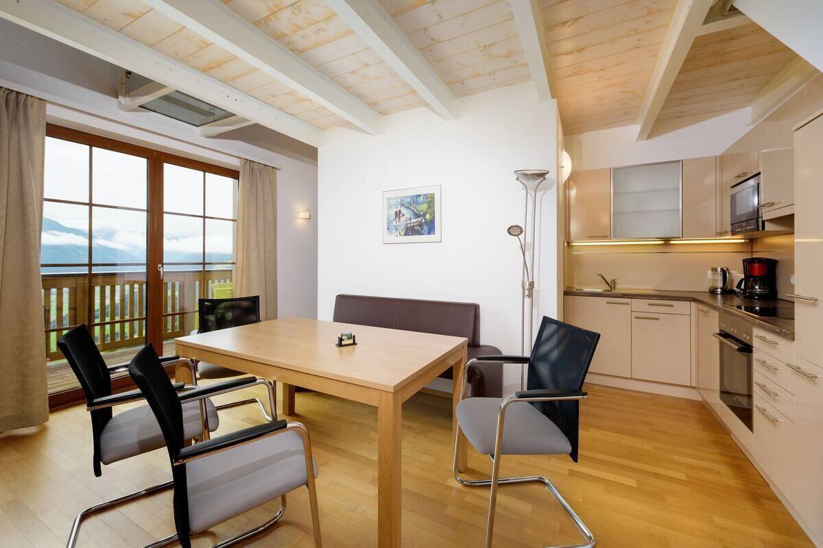 A room with a wooden table, chairs, a bench, a large window with a view, a painting on the wall, and a kitchenette.