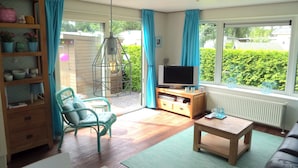 A living room with a television, a wooden cabinet, a green chair, a coffee table, and a view of greenery outside.