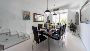 Et moderne spiseområde med en glasklædt trappe, et spisebord dækket med tallerkener, glas og en flaske, samt to hængende lysarmaturer.
