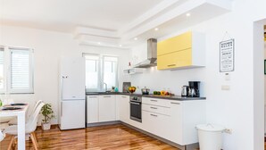 A modern kitchen with white cabinets, a yellow upper cabinet, and a stainless steel range hood. There is a dining area with a white table and chairs, and a potted plant. - Stunning home in Bilice (Bilice)