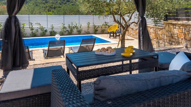 An outdoor seating area with a pool, wicker furniture, and a stone wall. - Stunning home in Bilice (Bilice)