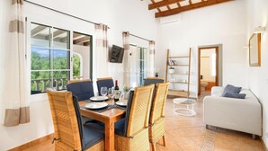 Zona pranzo con sedie in vimini, un tavolo in legno apparecchiato con piatti, bicchieri e una bottiglia di vino. Sullo sfondo, una TV a parete, un divano e una libreria.