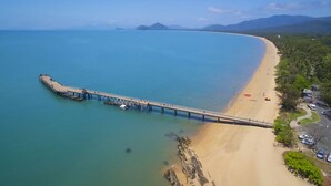 An aerial view of a coastal area with a long pier extending into the water, a sandy beach, and a road leading to a parking area.