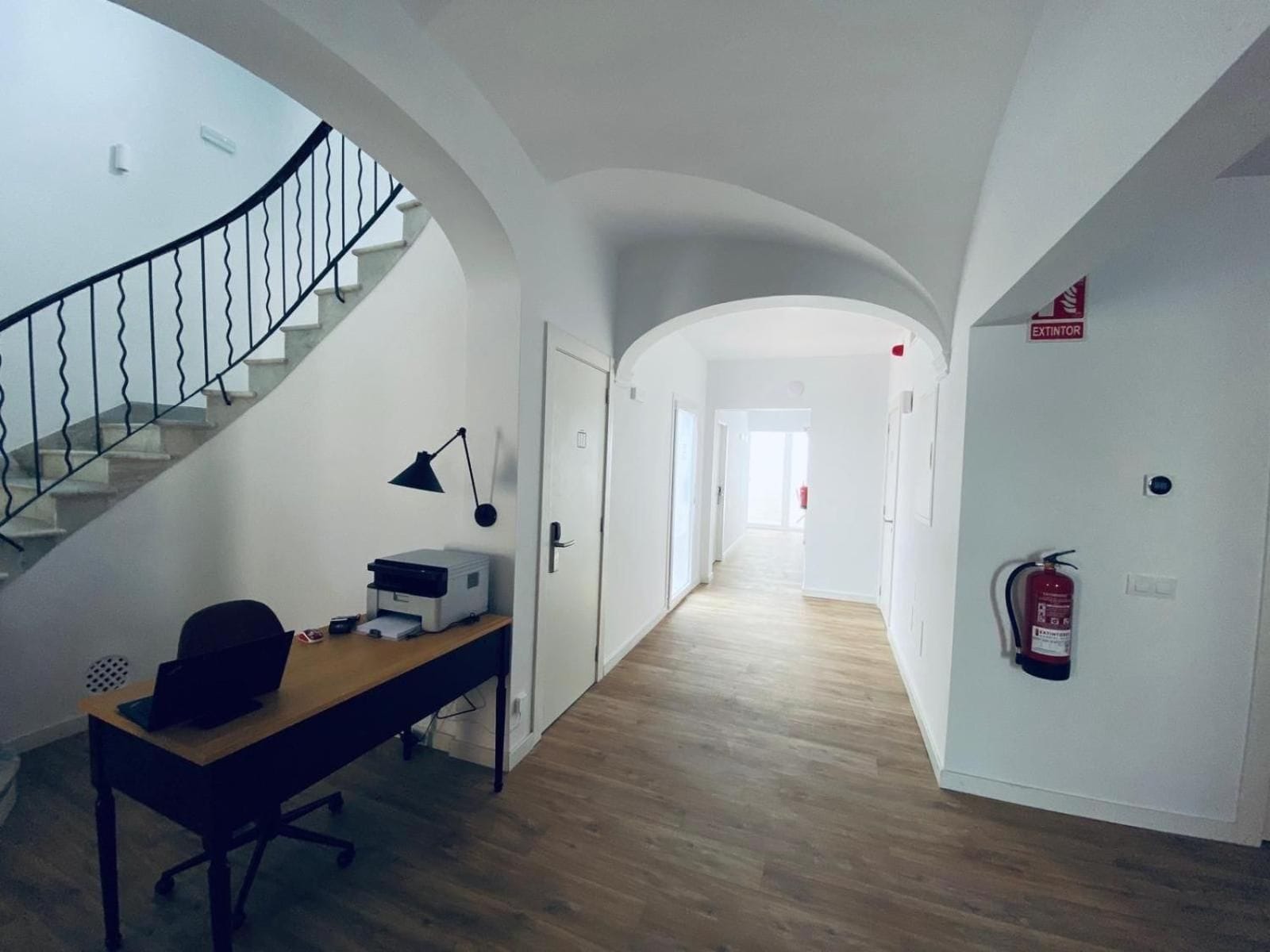 Un pasillo con una escalera a la izquierda, un escritorio con impresora y lámpara, y un extintor a la derecha.