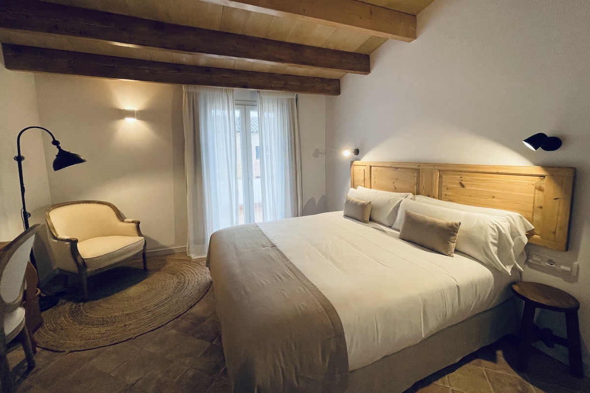 Um quarto com cabeceira de madeira, cama com roupa de cama branca, uma cadeira, um abajur e um pequeno banquinho.