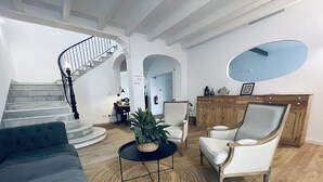 Una sala de estar con un sofá gris, una mesa redonda y una silla. Se ve una escalera con barandilla que lleva a otra habitación.