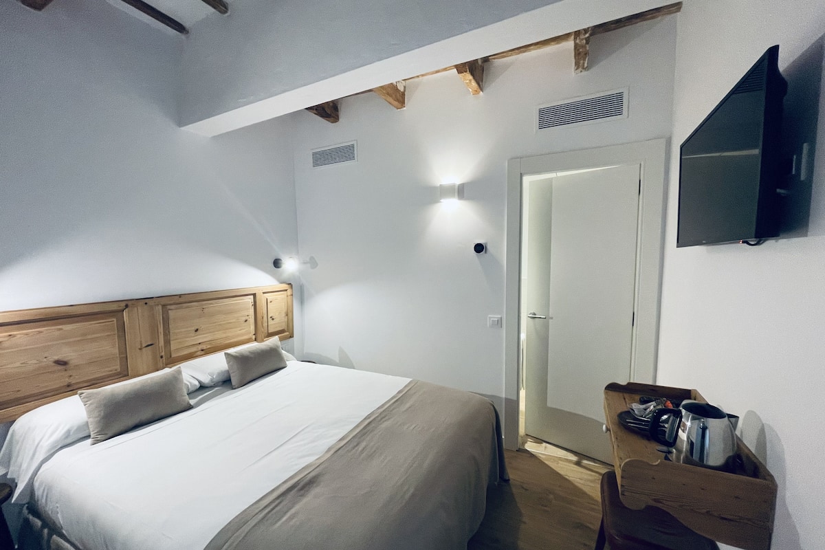 Quarto com cabeceira de madeira, cama com roupa de cama branca, mesa de cabeceira com chaleira e televisão fixada na parede.