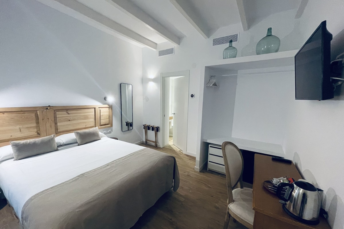 Quarto com uma cama grande, cabeceira de madeira, mesa de cabeceira, uma cadeira, escrivaninha com uma chaleira e TV na parede.