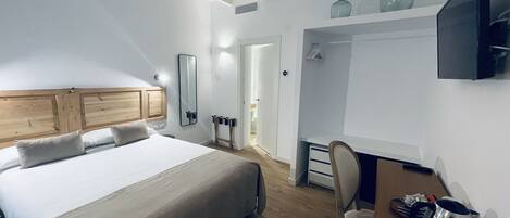Un dormitorio con una cama grande, cabecera de madera, mesita de noche, una silla, escritorio con tetera y televisor montado en la pared.