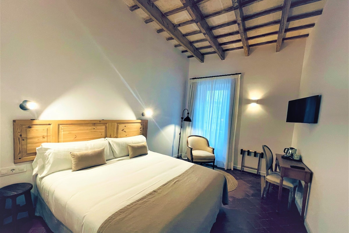 Quarto com cabeceira de madeira, cama com roupa de cama branca, uma cadeira, escrivaninha com televisão de tela plana e janela com cortinas.