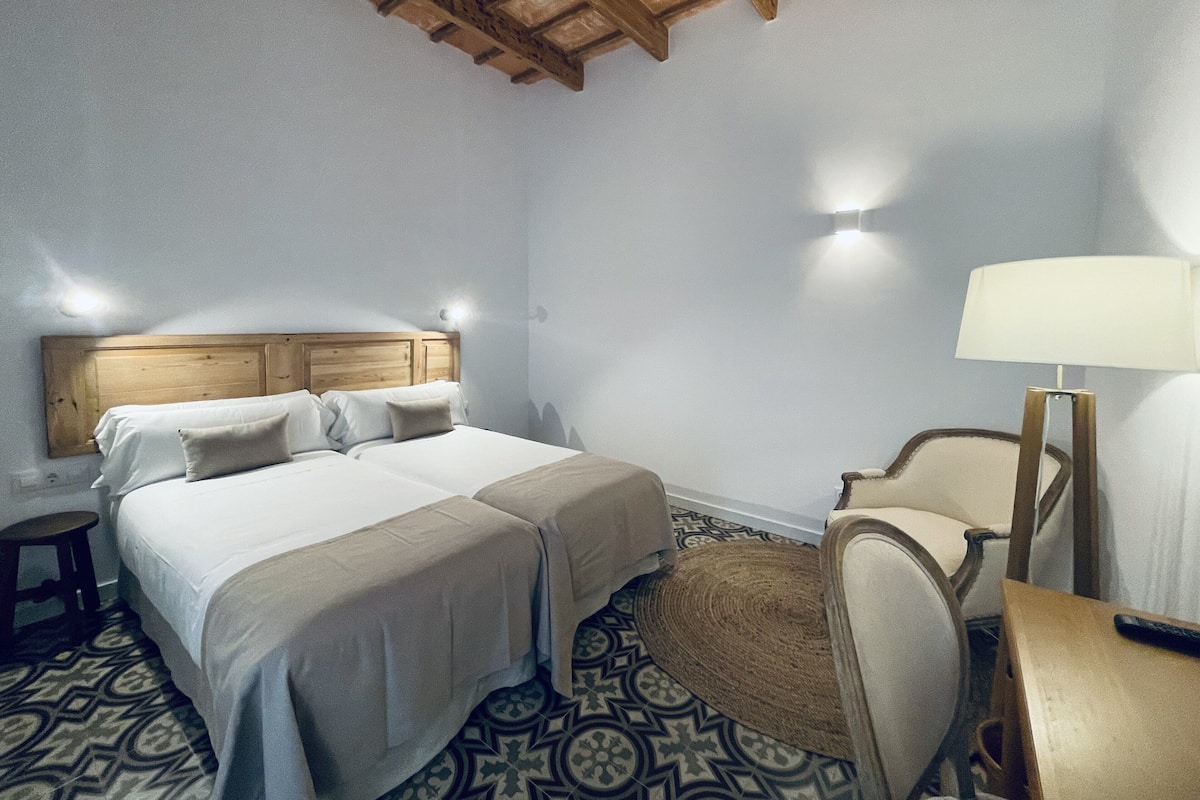 Quarto de hotel com teto de madeira, duas camas com roupa de cama branca, uma mesa redonda pequena, uma cadeira e um abajur.