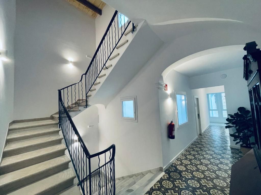 Una escalera con barandilla negra que lleva a un rellano, un suelo con diseño y un pasillo con una puerta y una planta.