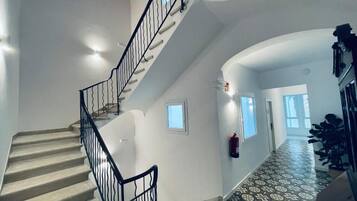 Una escalera con barandilla negra que lleva a un rellano, un suelo con diseño y un pasillo con una puerta y una planta.
