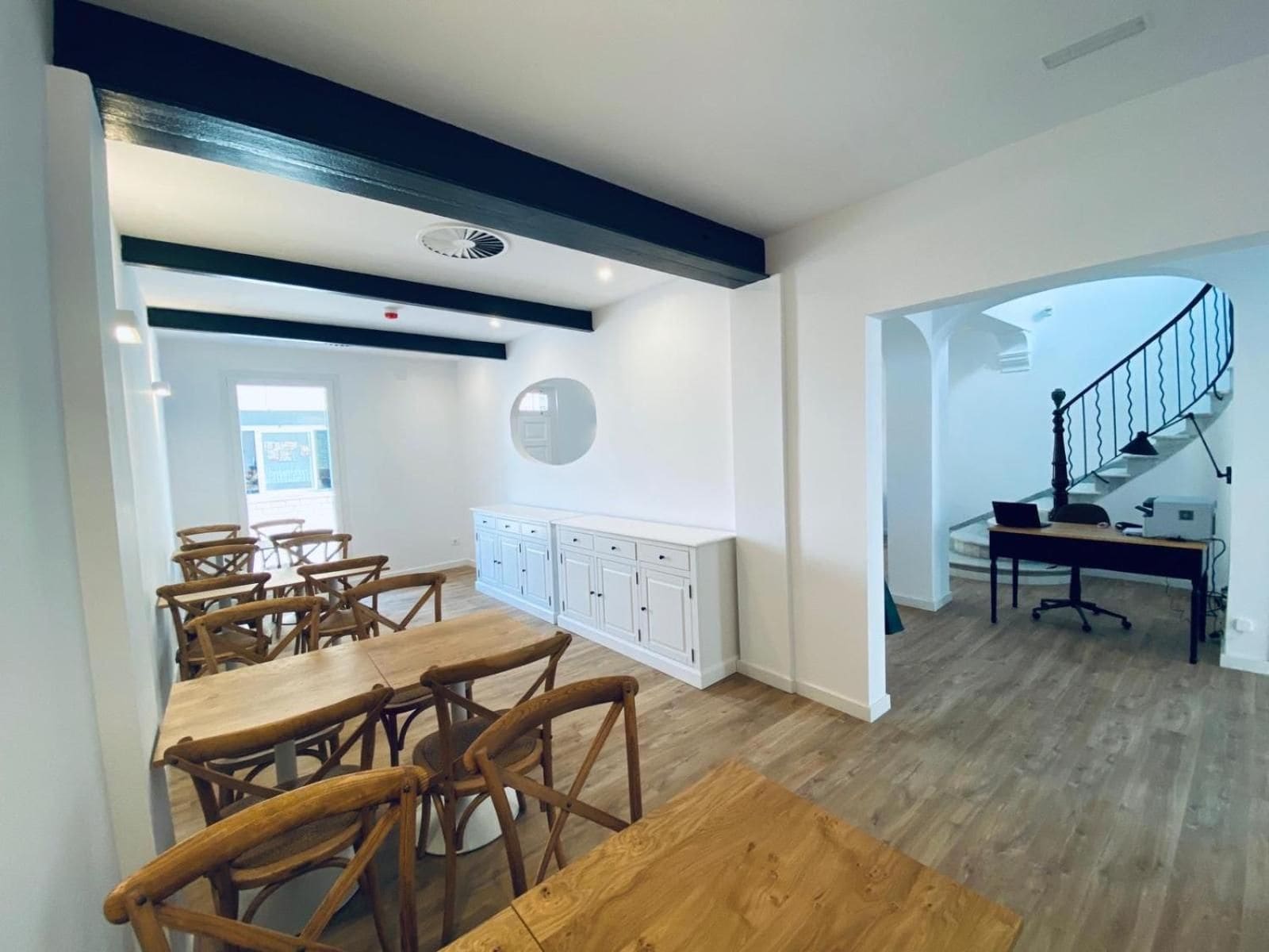 A spacious dining area with wooden tables and chairs, a white cabinet with drawers, and a staircase leading to an upper level.
