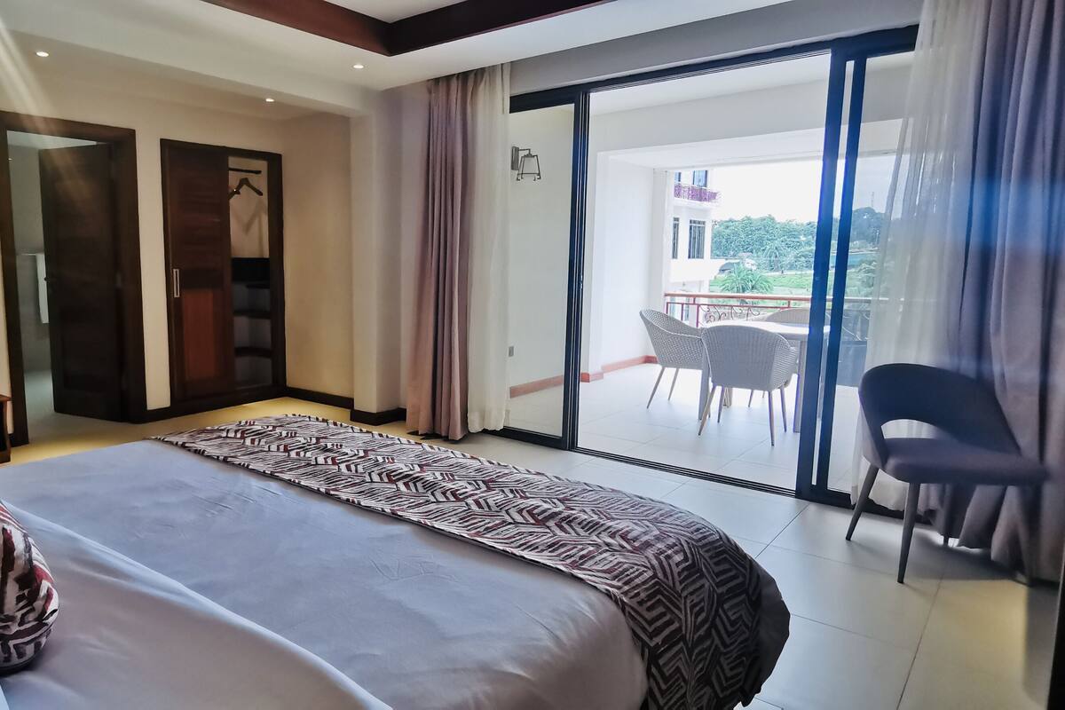 A hotel room with a bed, a wardrobe, a balcony with chairs, and a view of the outside.